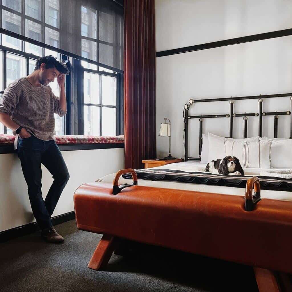 Man standing in a room with a dog on a bed.
