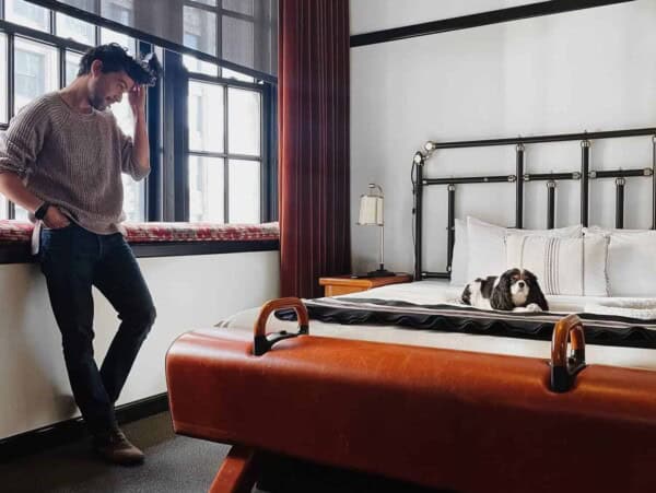Man standing in a room with a dog on a bed.