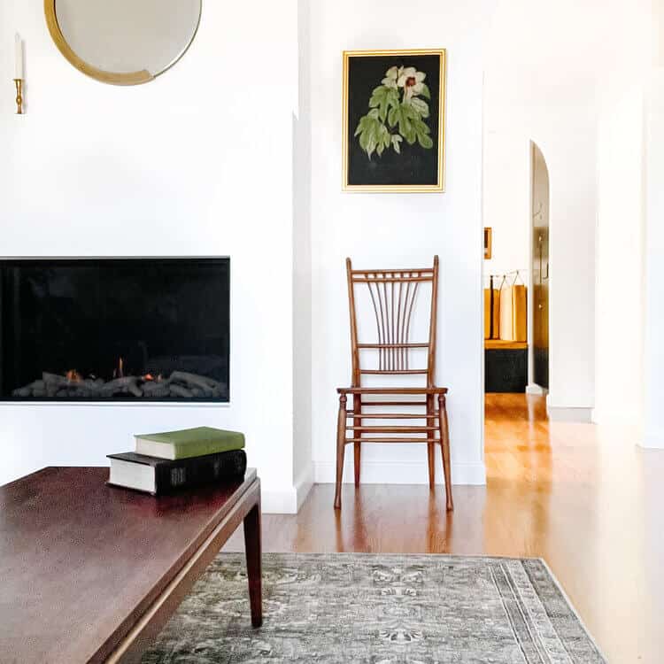 Plant picture above a chair next to a fireplace below a mirror.