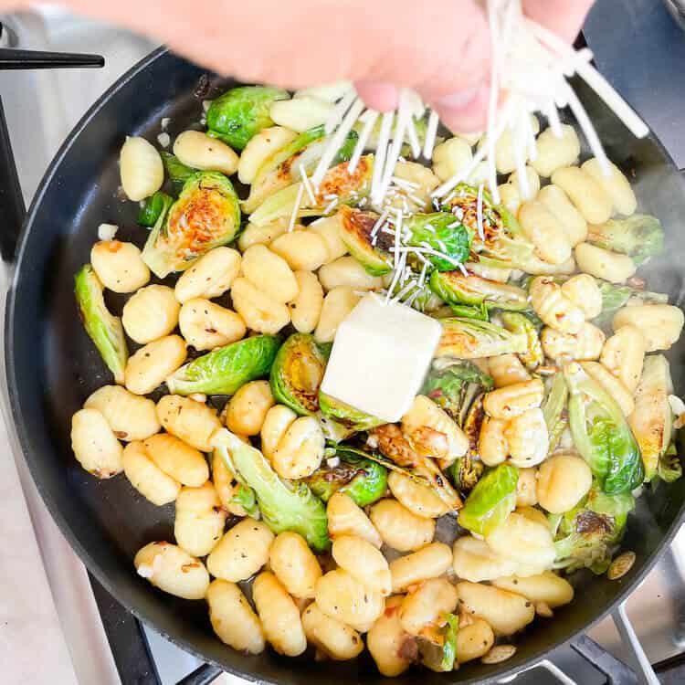 Person sprinkling cheese on a pan of brussels sprouts, gnocchi, and butter.