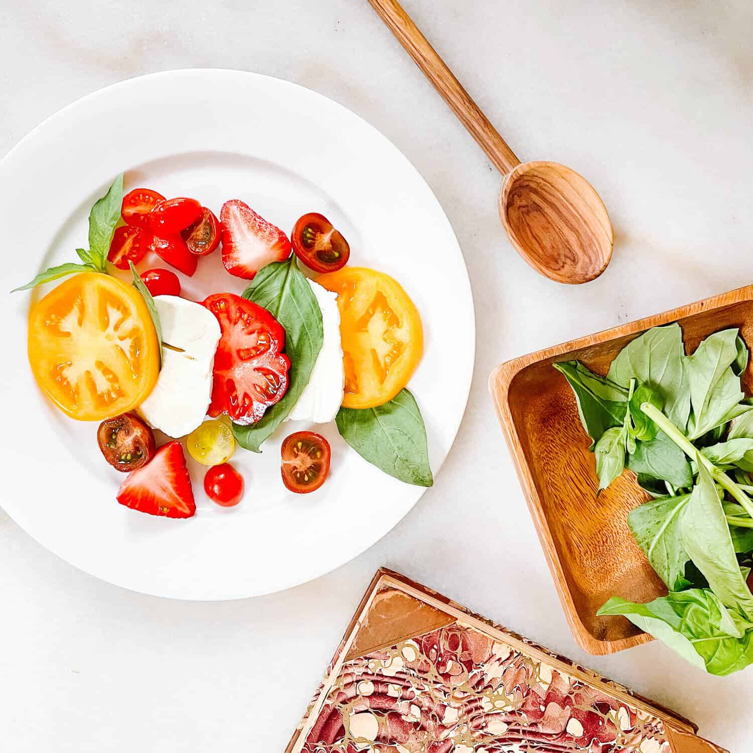 strawberry caprese salad on a table with fresh basil and tomatoes on a whitSe plate. 