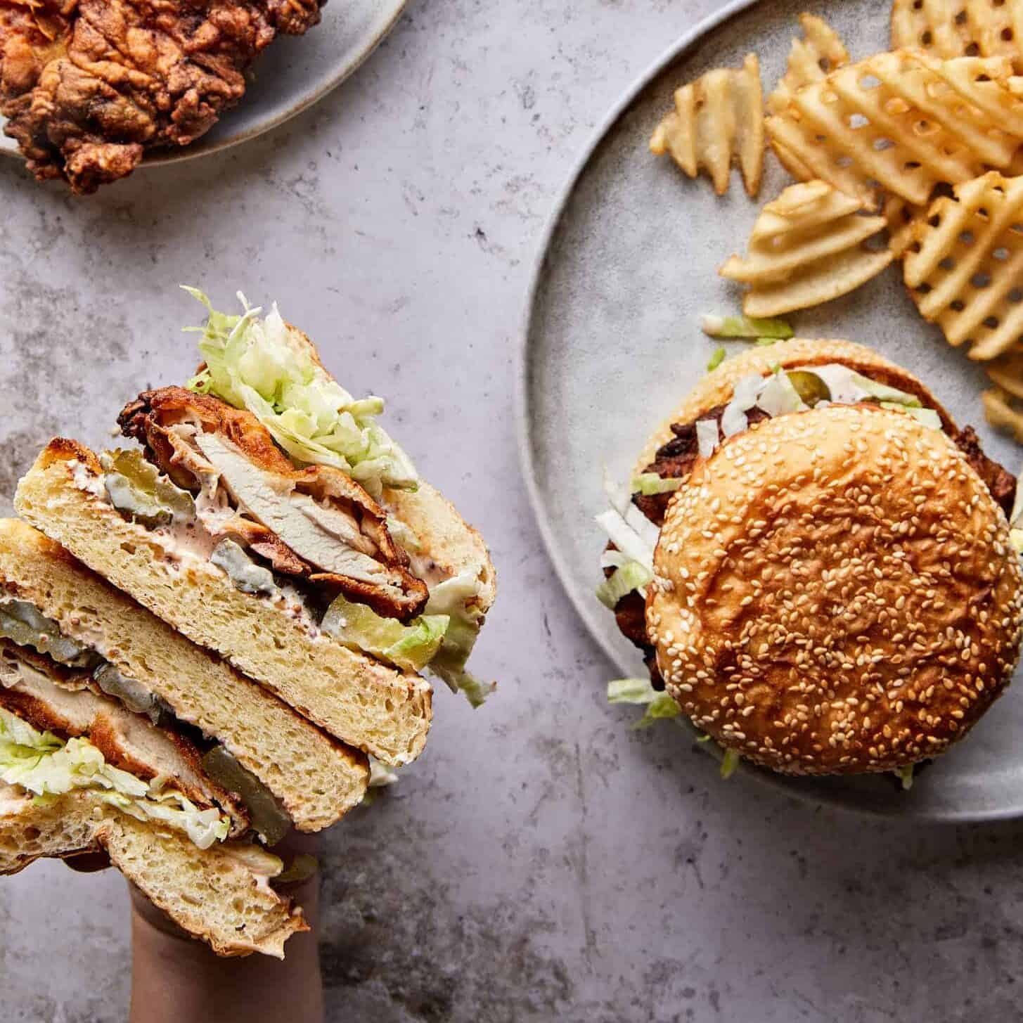 The best fried chicken sandwich recipe you'll ever try—golden, crispy chicken on a buttery brioche bun with tangy pickles, spicy oil, and a kick of something sweet being held over a plate of waffle fries.
