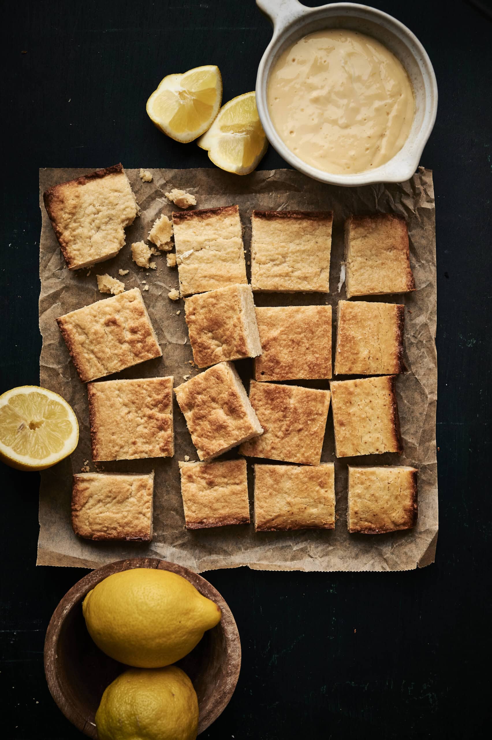 Moist lemon cookie bars with cream cheese frsoting ina pot cut into clean, even squares. on a black table and parchment paper. 
