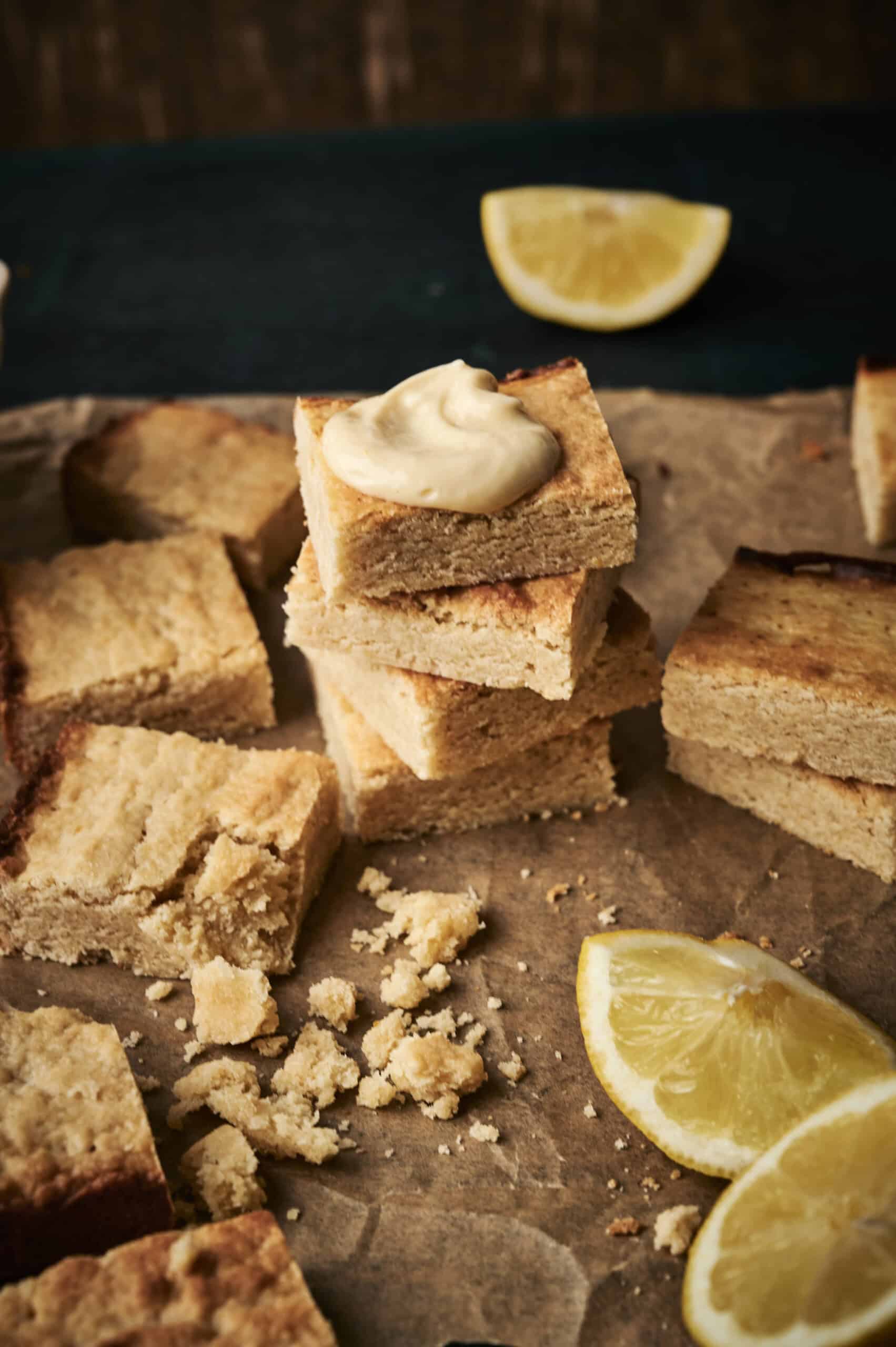 Freshly baked lemon cookie bars with cream cheese frosting on top, resting on parchment with sliced lemon wedges and crumbles. 