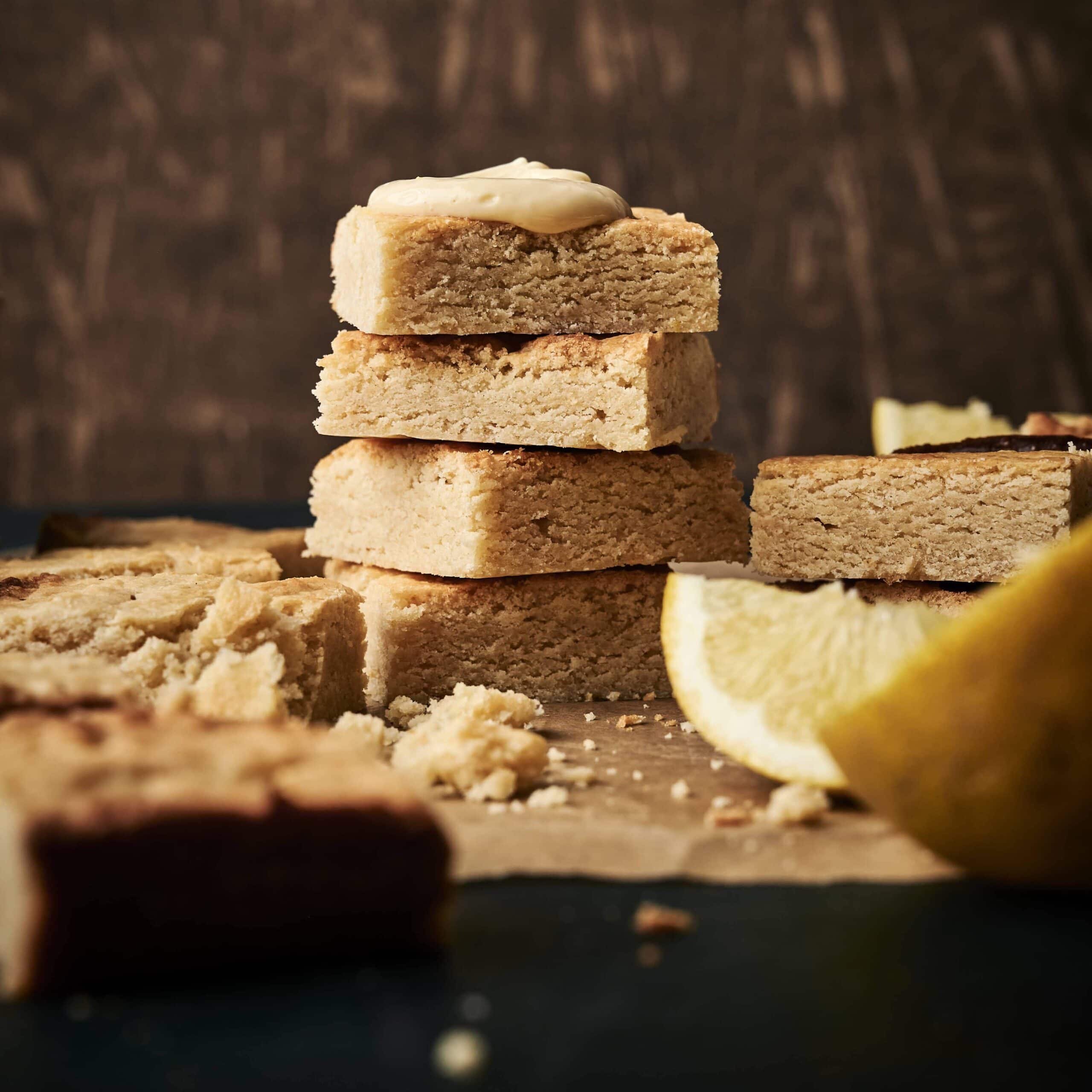 A batch of lemon cookie bars with cream cheese frosting on top, stacked high, cooling next to fresh slices of lemon.