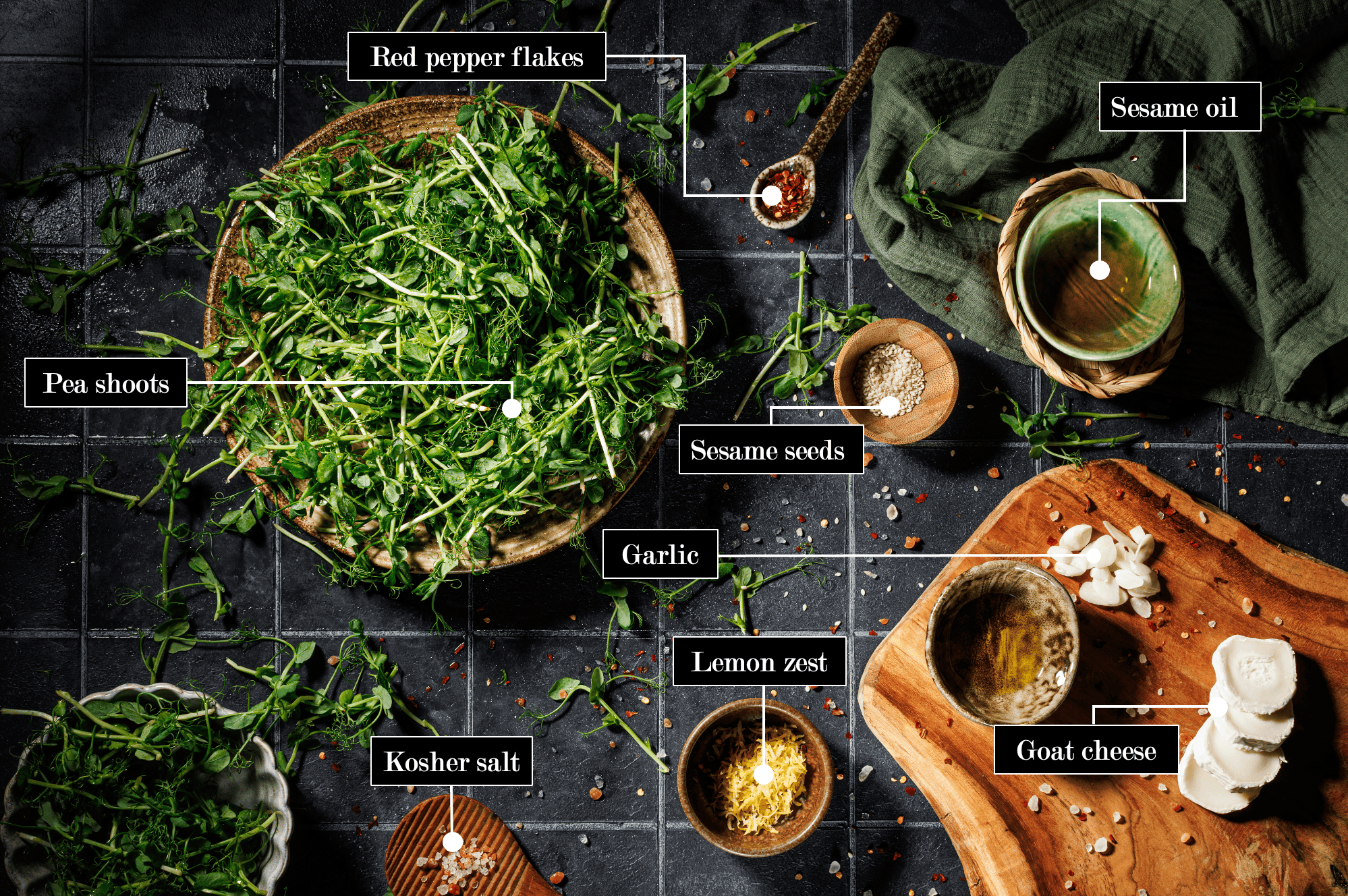 Pea shoots ingredients on a black stone table with spilt red flaks featuring sesame oil, garlic, fresh spea shoots, and garlic to name a few