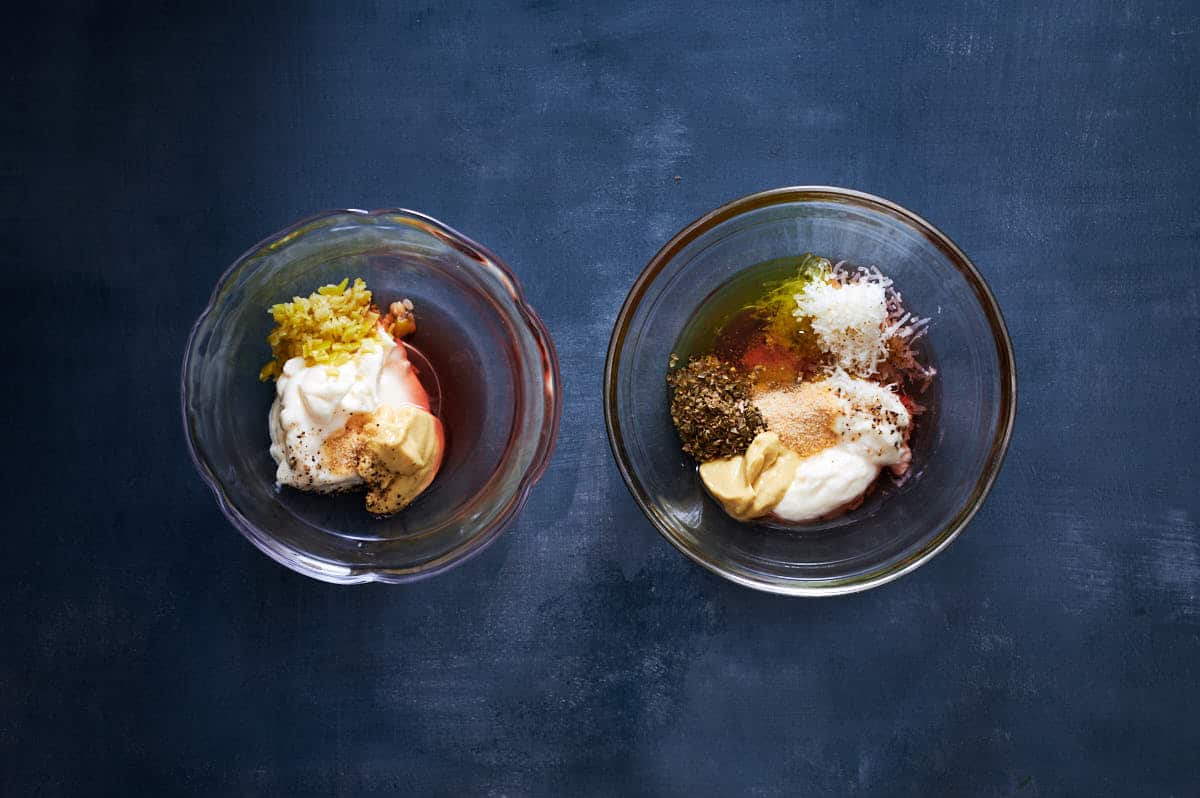 Two ramekins full of wet ingredients for both a dressing and aioli on a blue table.