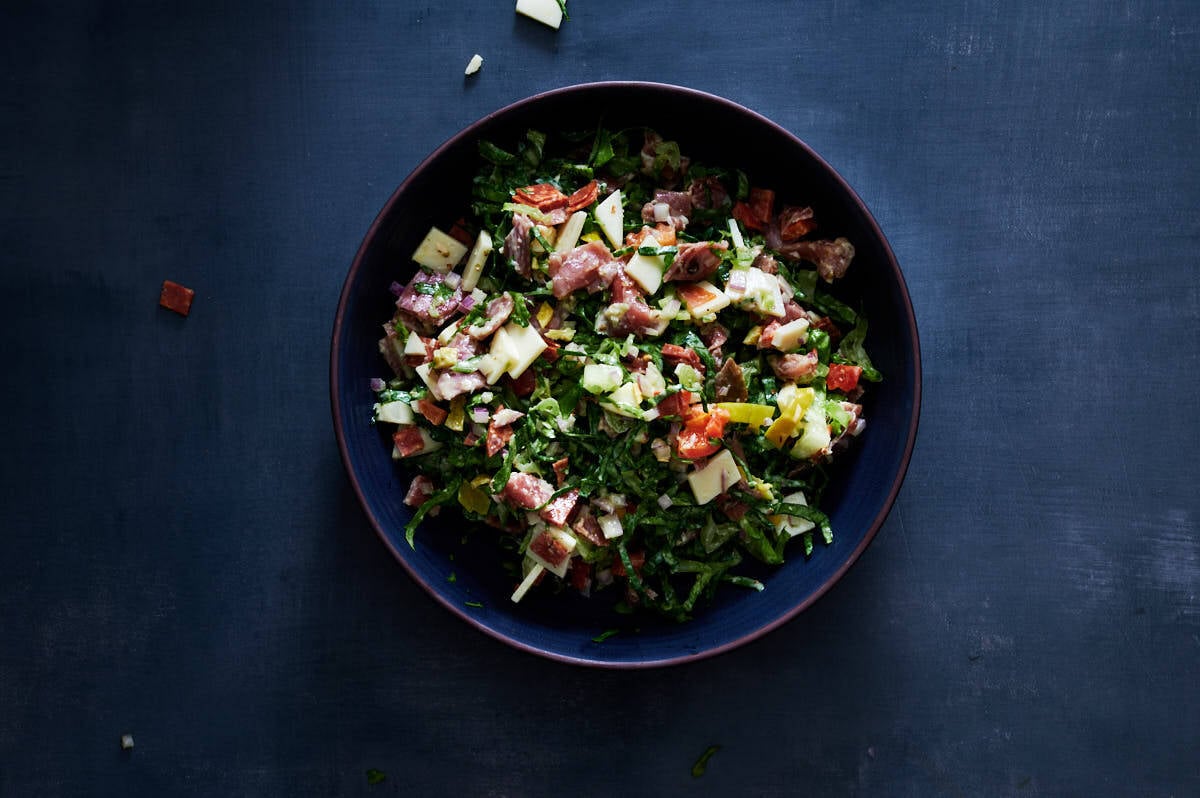 Mixing the chopped meat, cheese, lettuce, and peppers in a large glass bowl with creamy Italian dressing.
