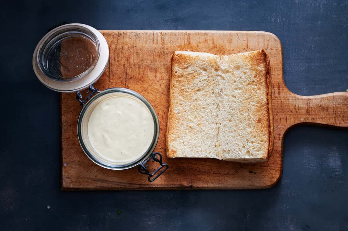 chibata bread cut open faced with aioli ready to spread on a wooden cutting board on top of a blue table.