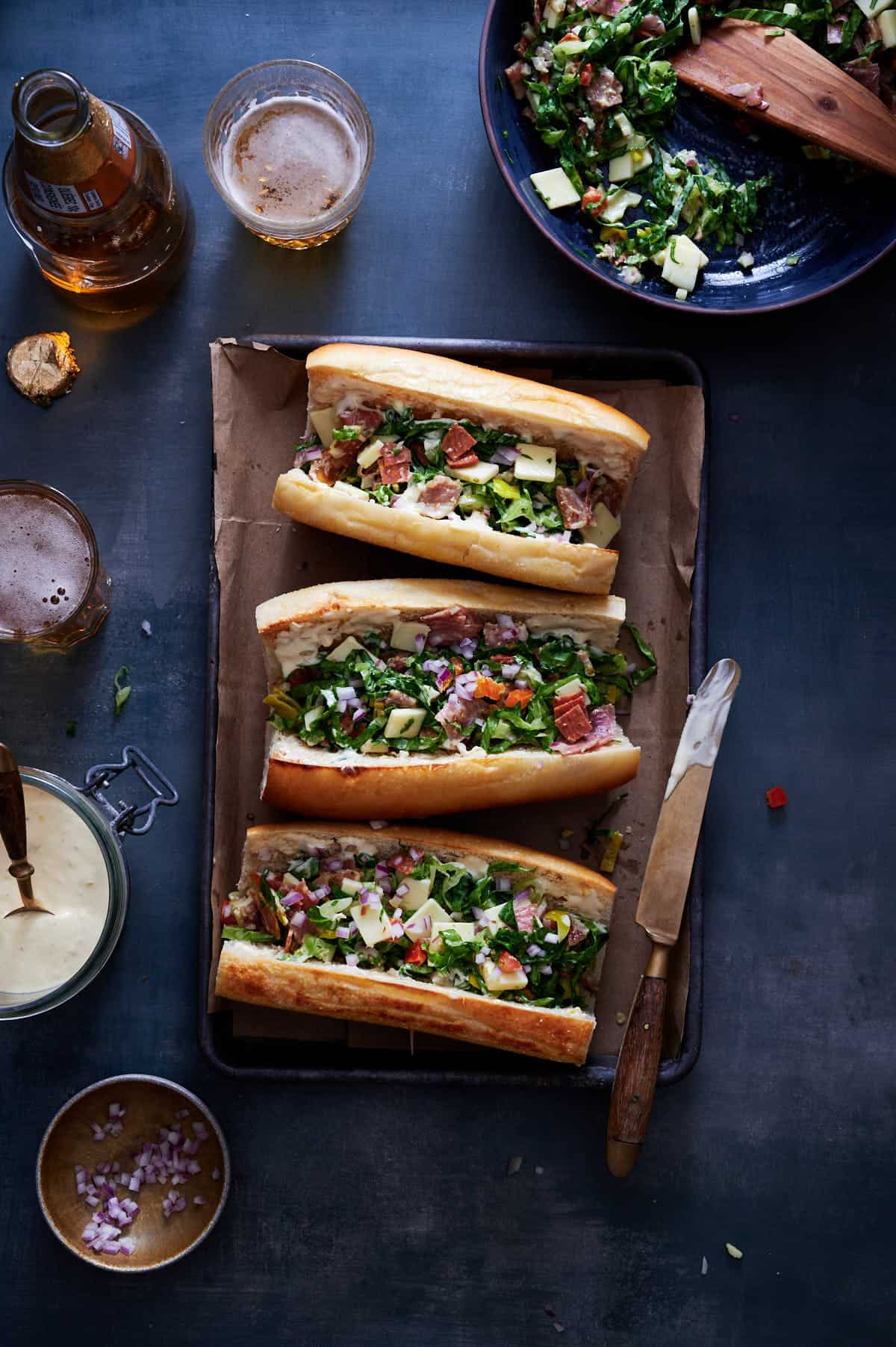 Chopped Italian sandwich on toasted ciabatta, layered with meats, provolone, lettuce, and balsamic glaze on a blue table with a poured beer.

