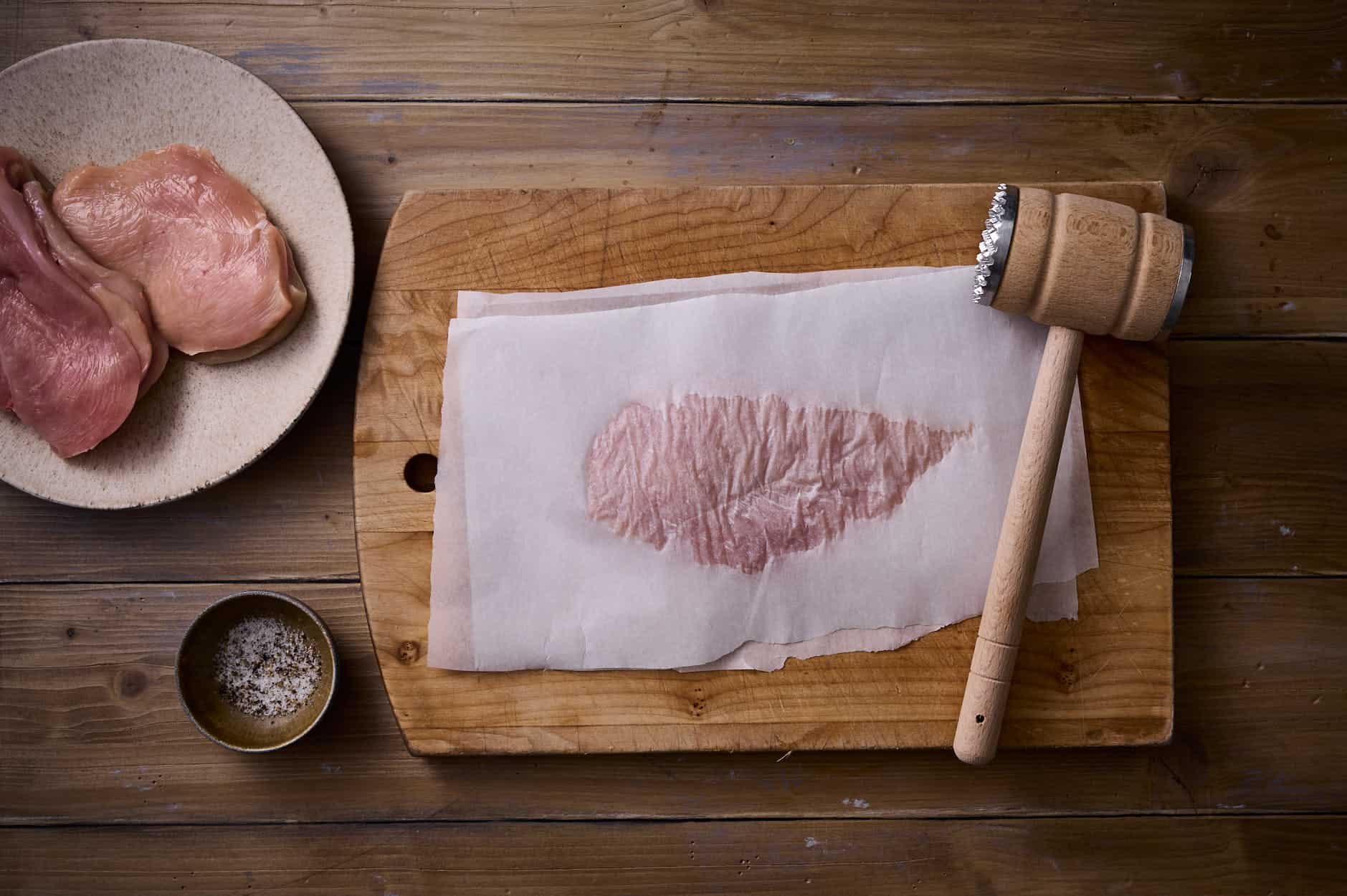 Picatta milanese being pounded thin between parchment paper with a mallet on a wooden table. 