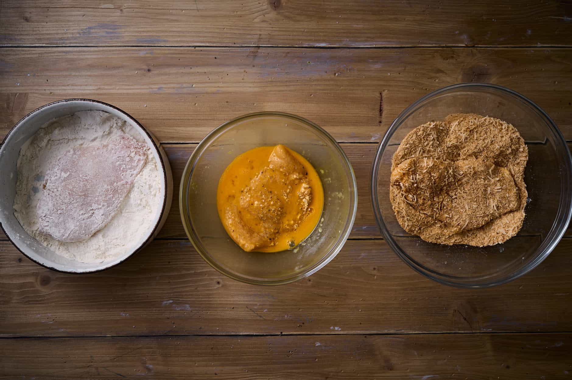 dredging ingredients on a wooden table for Picatta Milanese, including flour, beaten egg, and Italian bread crumbs. 