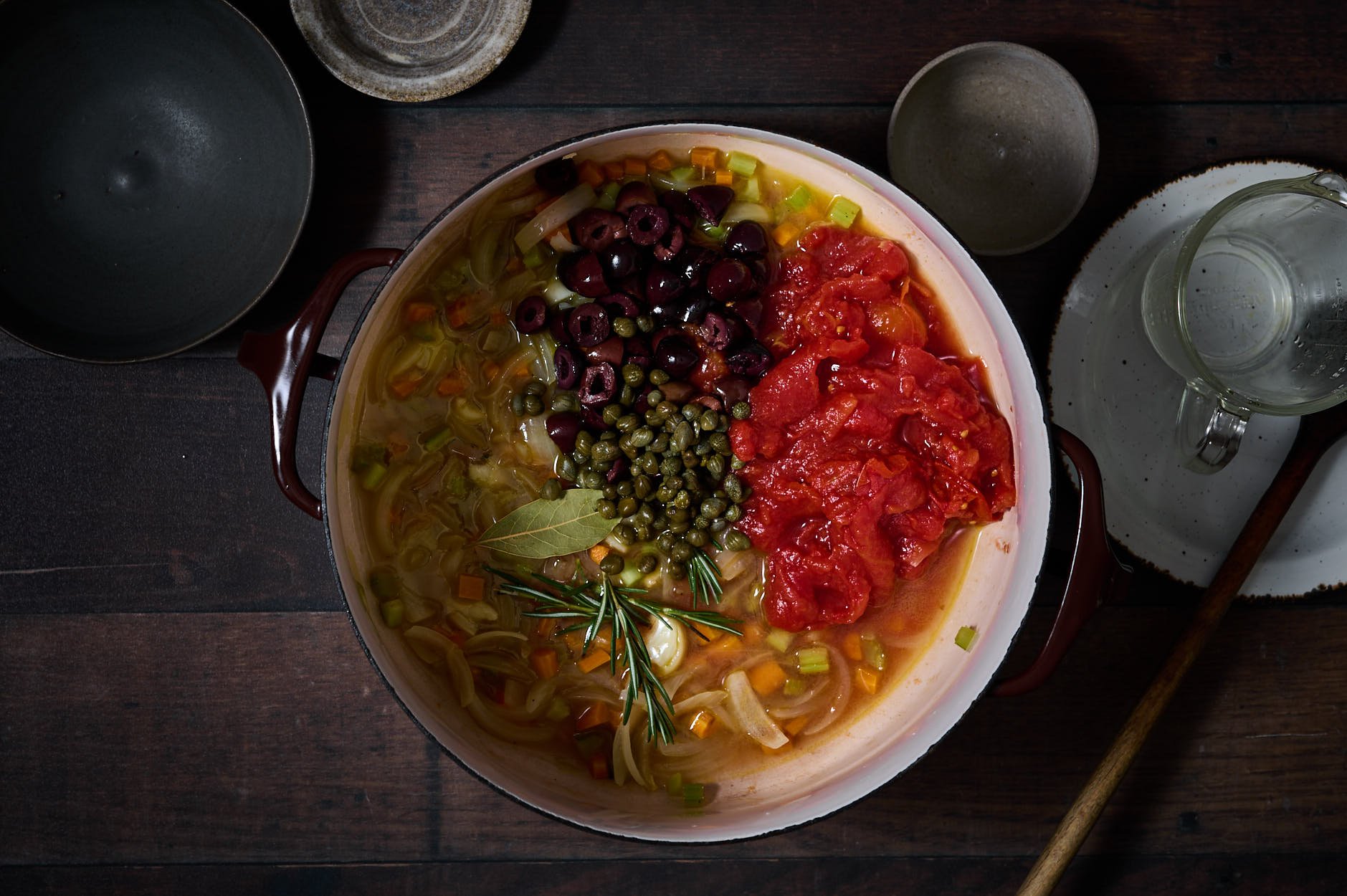 a crockpot is simmering full of white wine, diced vegetables, and the addition of crushed tomatoes, olives, capers, and rosemary on a rustic table.