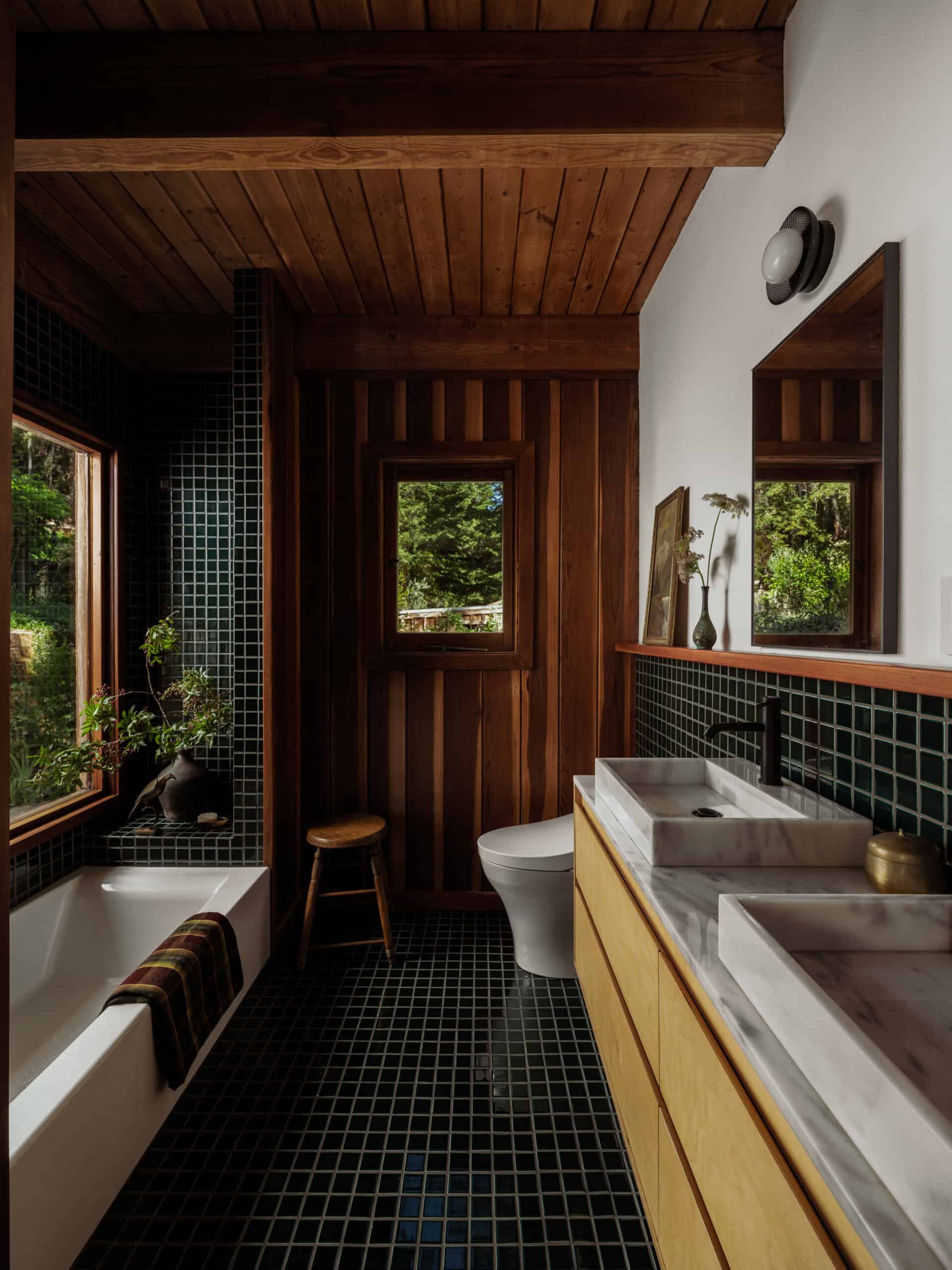 The final aftter image of a mody bath with fresh green shiny tiles. The vanity is tan with marble tops and matte black faucets. There are new medicine cabinets with black trim and a vintage stall with flowers and vases. 