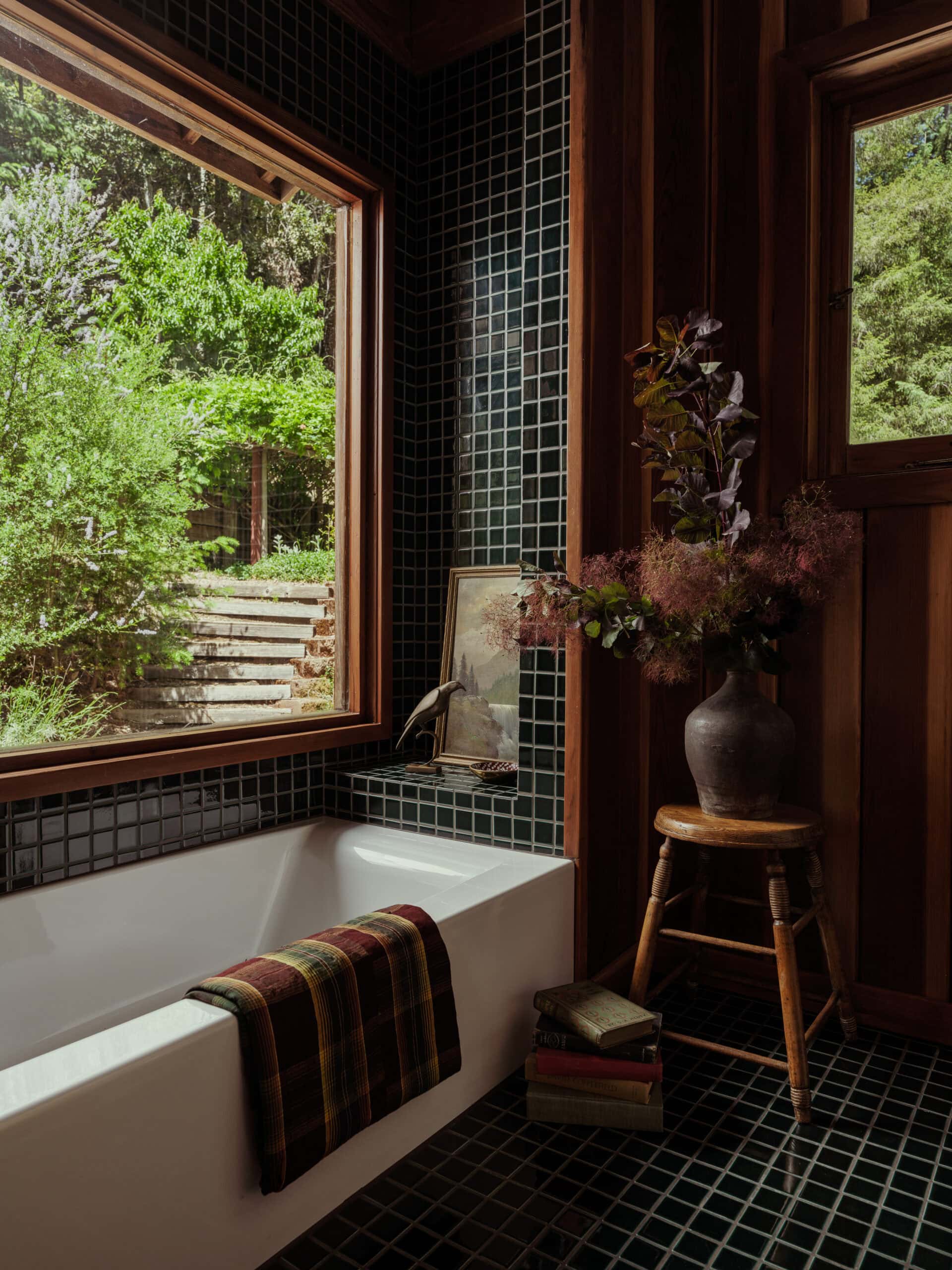 The after moody bath featuring evergreen 2x2 glossy tiles, refreshed redwood and a giant picture frame window with greenery on the outside and a vintage stool with stacked books. Looks amazing.