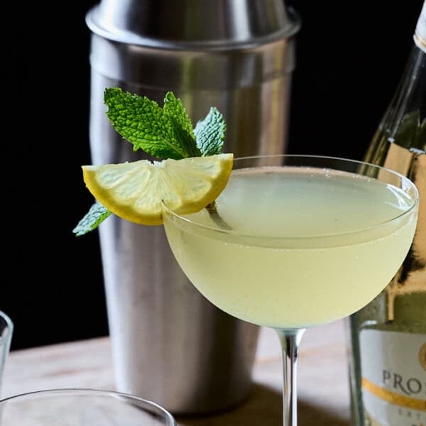 A Lemoncello cocktail in a up glass with fress mint and lemon onto of a wooden table with an opened bottle of prosecco.