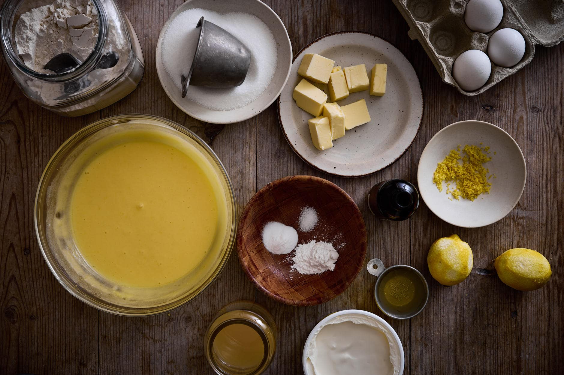 Ingredients for lemon poppy seed cake arranged on a wooden table surface; flour, eggs, lemon juice, butter and zest with sour cream and cream cheese etc. 