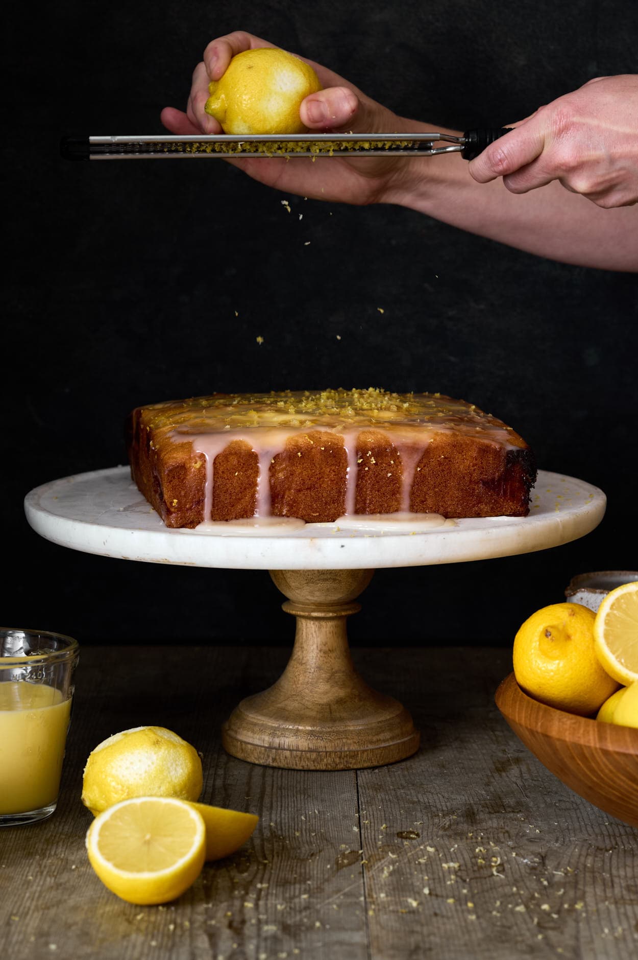 Lemon and poppy seed cake loaf being zested with fresh glaze on a marble cake tray a top a wooden table with sliced lemons. 