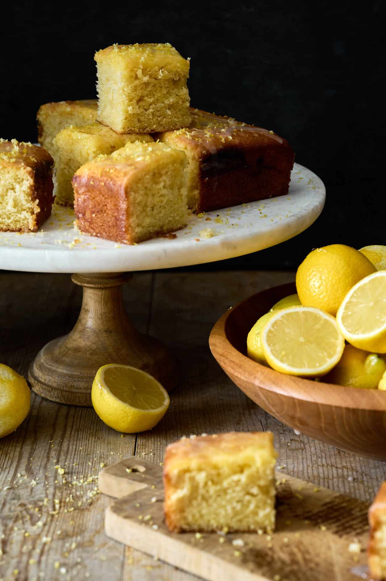 Lemon curd cake on a farm table with a marble tray next to a wooden bowl of fresh-cut lemons.