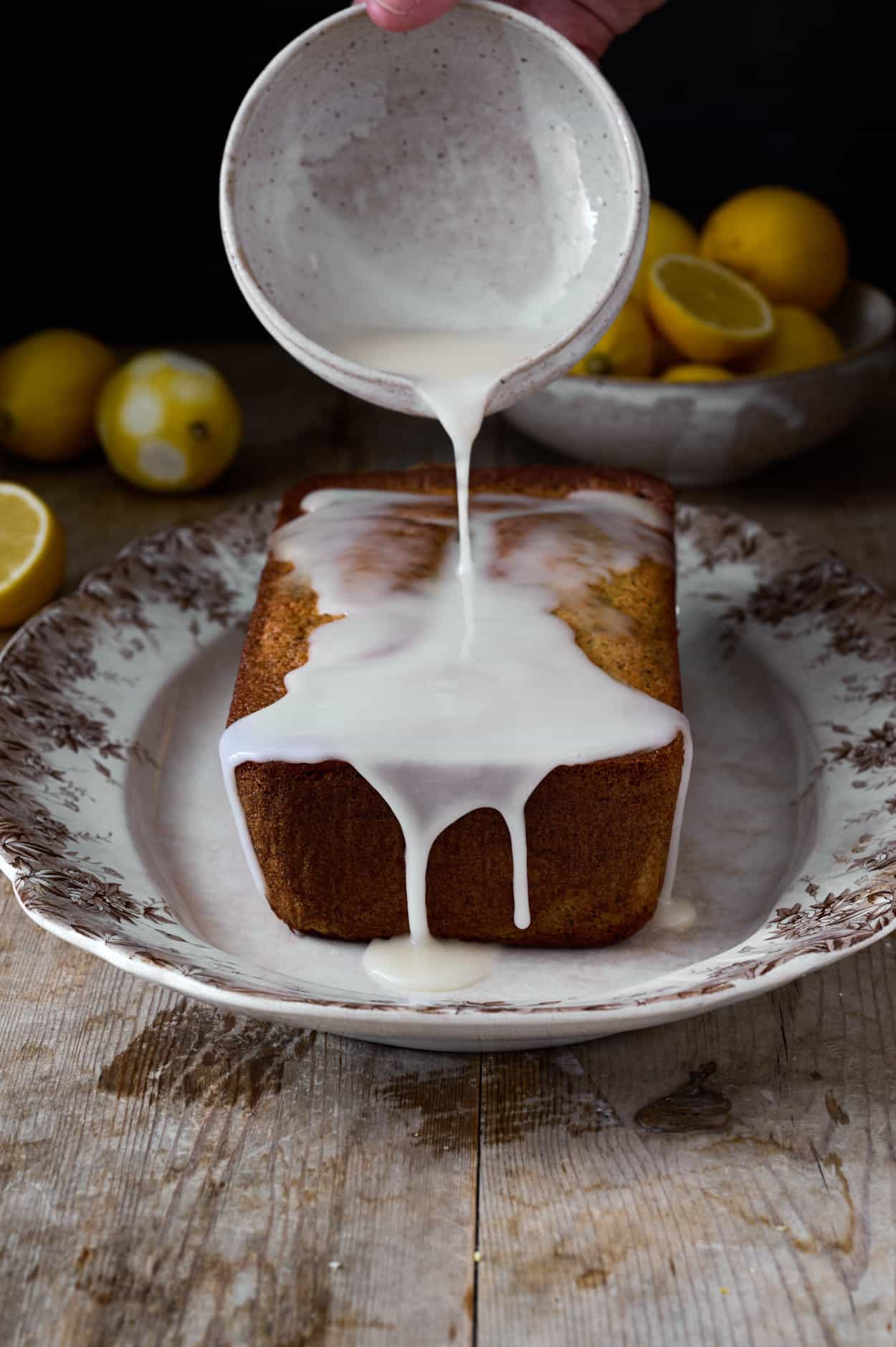 lemon poppy seed cake on a wooden table with vintage china being glazed from a bowl looking delicious and moist. 