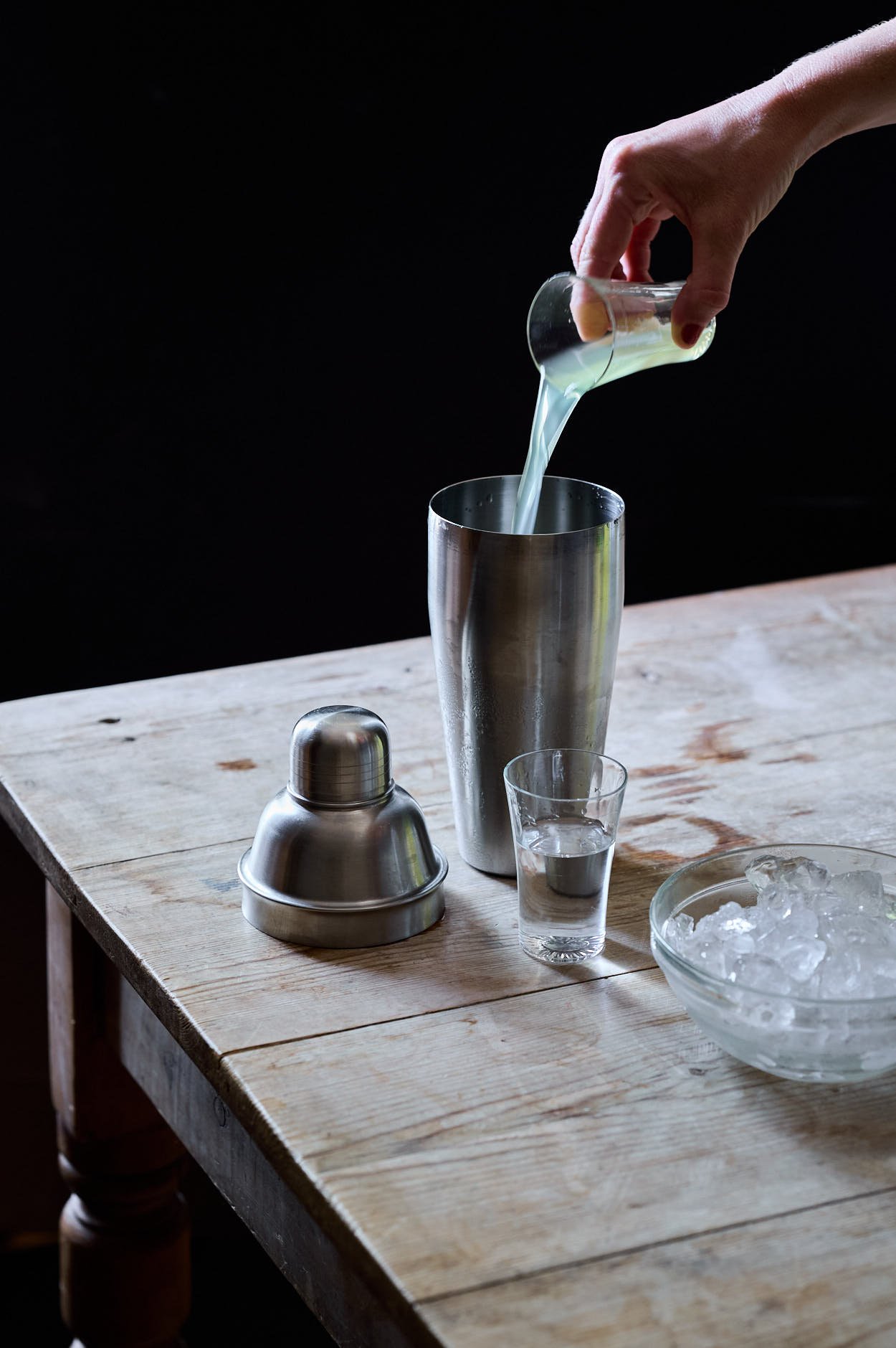 Pouring the lemon liquor into a chilled shaker on a wooden table with vodka and ice cubes. 