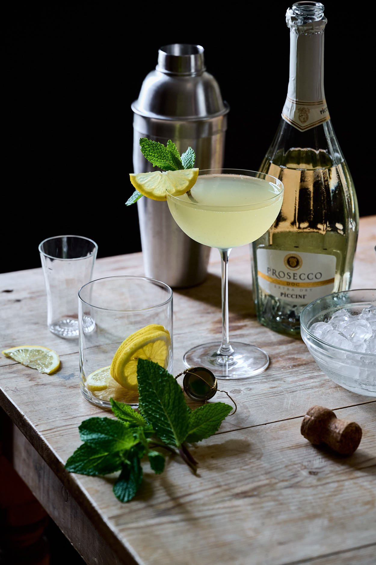 A Lemoncello cocktail in a up glass with fress mint and lemon onto of a wooden table with an opened bottle of prosecco.