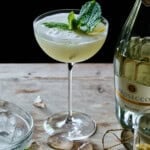 a fresh lemoncello cocktail freshly pouree in an up glass with a mint garnish, on a wooden table with an open bottle of prosecco.