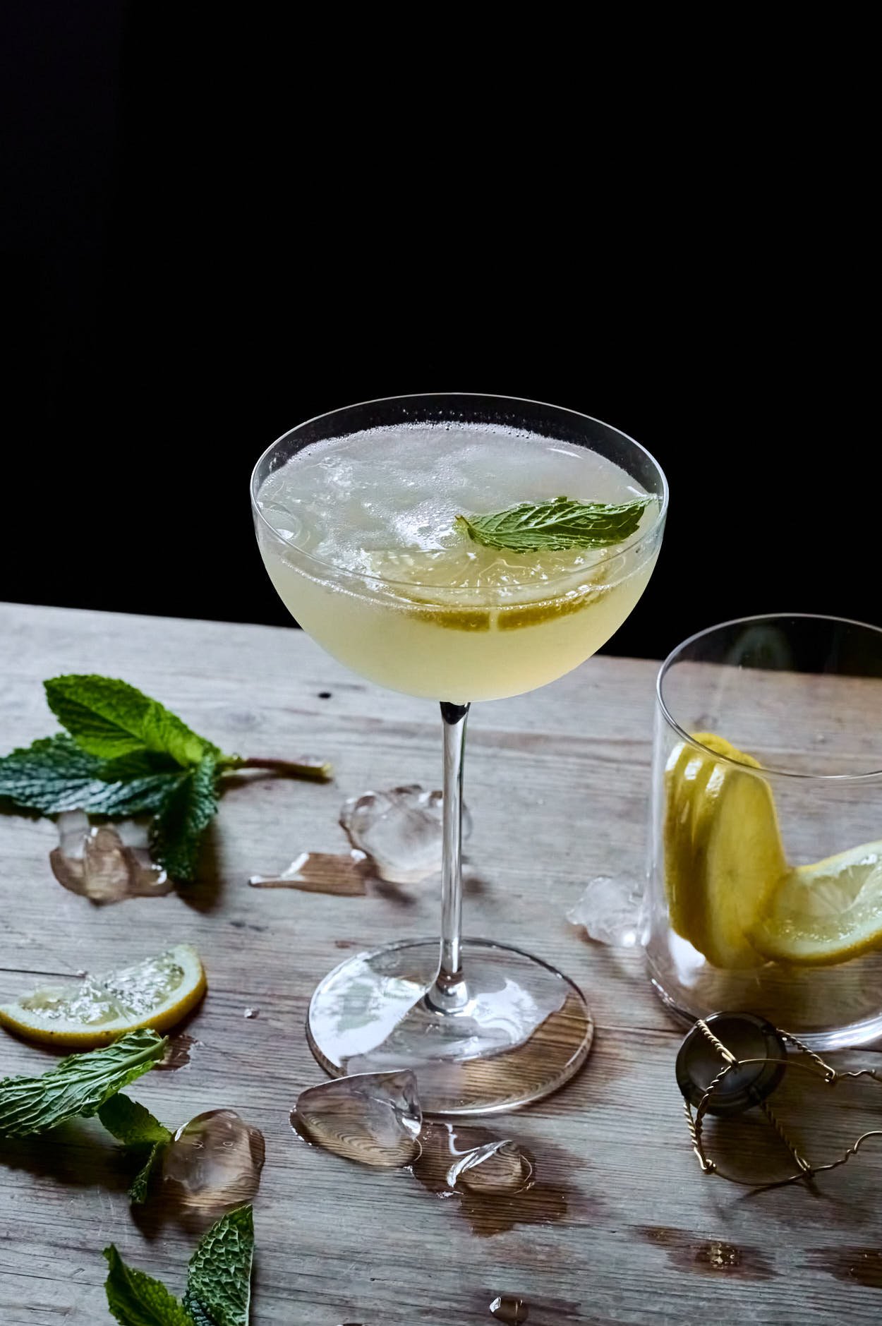 a Lemoncello cocktail with all the ingredients around it, including a bottle of prescoo, mint, lemon, and a shaker on top of a wooden table and a black backdrop. 