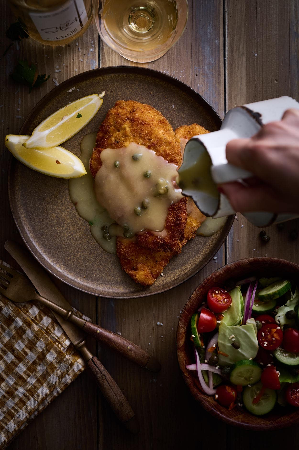 Pouring zesty lemon caper sauce onto Picatta milanese served on a brown terracotta plate with a glass of white wine and a salad. 