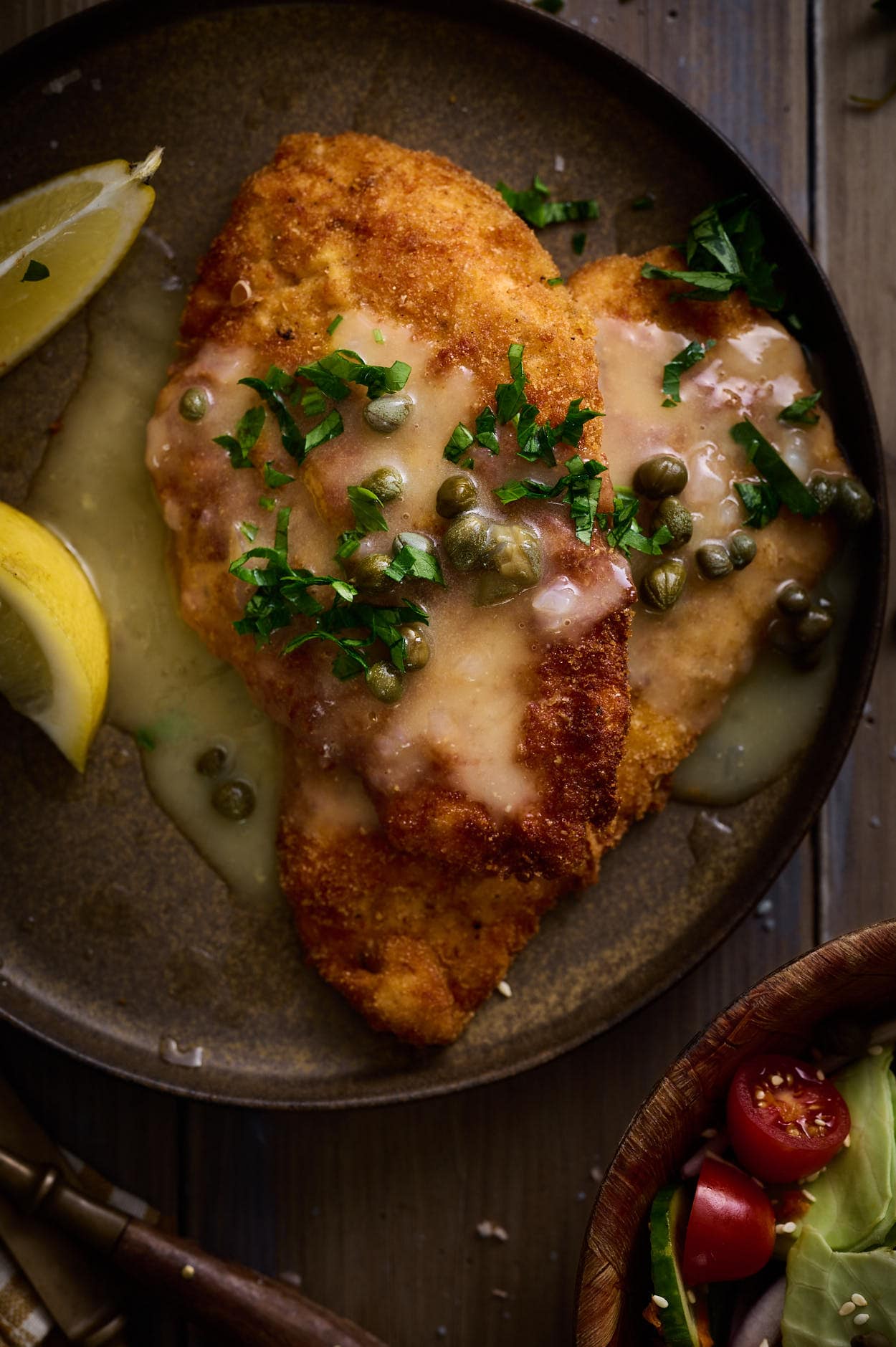 Picatta milanese served on a brown terracotta plate with shimmering lemon caper sauce, fresh lemon edges, and parsley. 