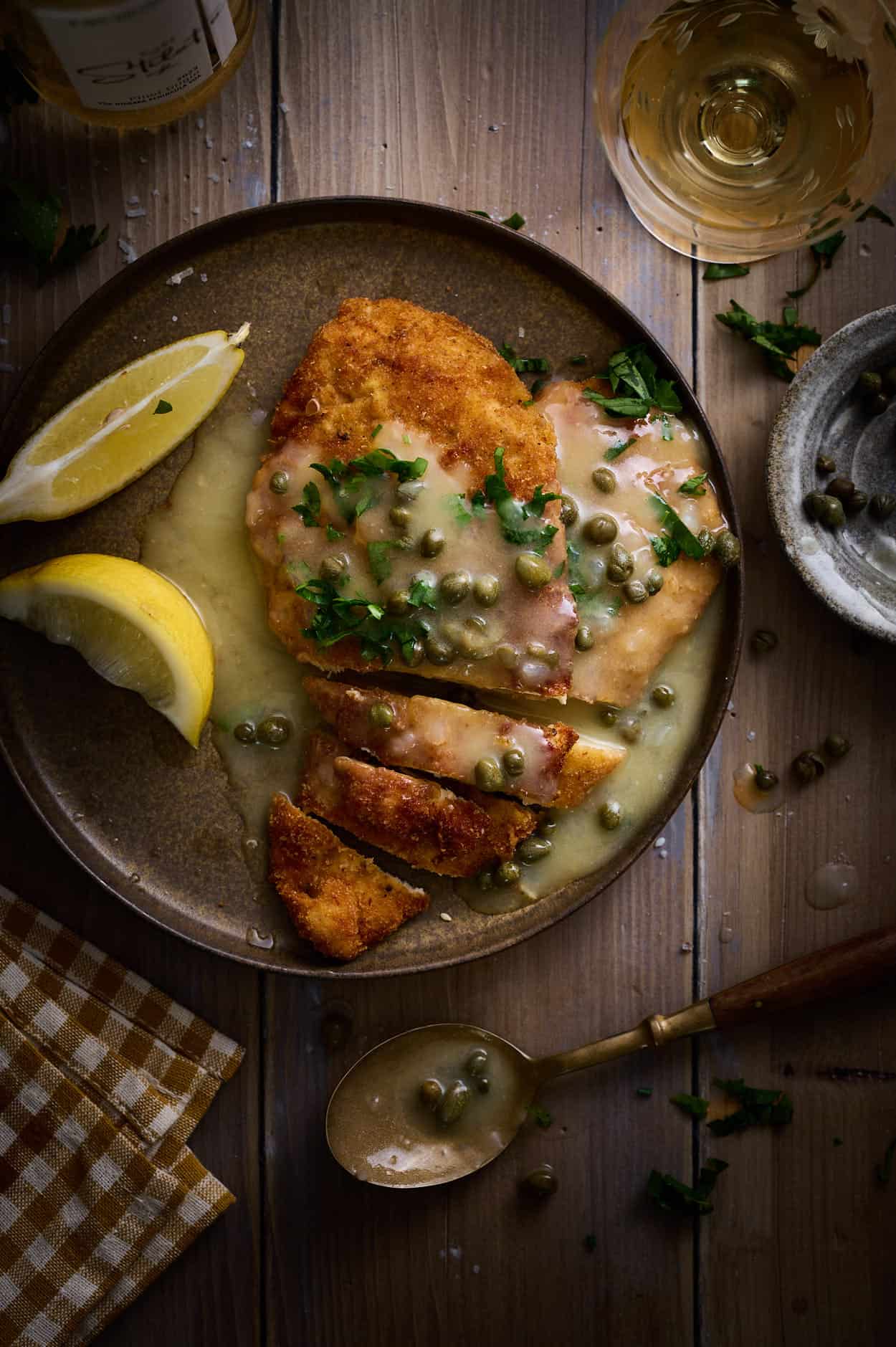 Zety lemon sauce covered picatta Milanese cutlet on a wooden table with white wine, lemon wedges, and sliced to eat .