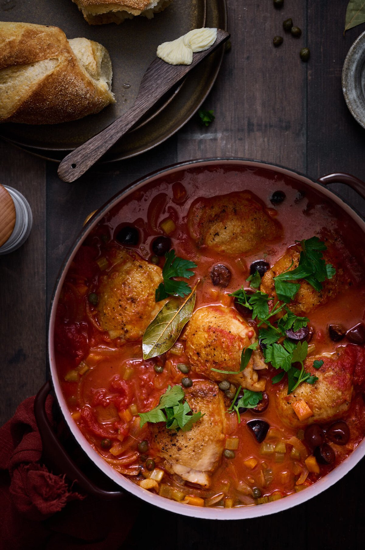 A Rustic Pollo alla Cacciatora Recipe on a distressed brown table in bubbling tomato sauce, with fresh parsley garnish and chicken thighs. 