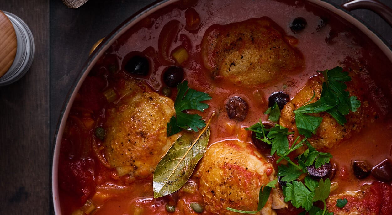 Pollo all Cacciatora on a table in bubbling red tomato sauce, with seared chicken thighs, fresh green herbs, and crusty bread and butter.