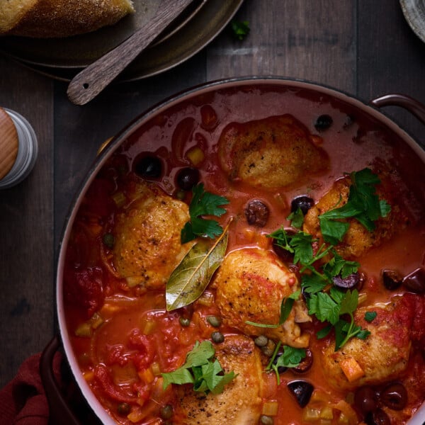Pollo all Cacciatora on a table in bubbling red tomato sauce, with seared chicken thighs, fresh green herbs, and crusty bread and butter.