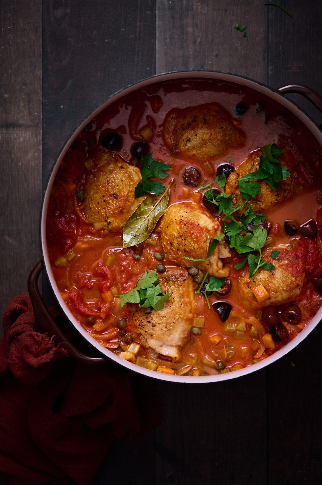Pollo all cacciatora with red tomato sauce and fresh green herbs in a dutch oven on a rustic table. 