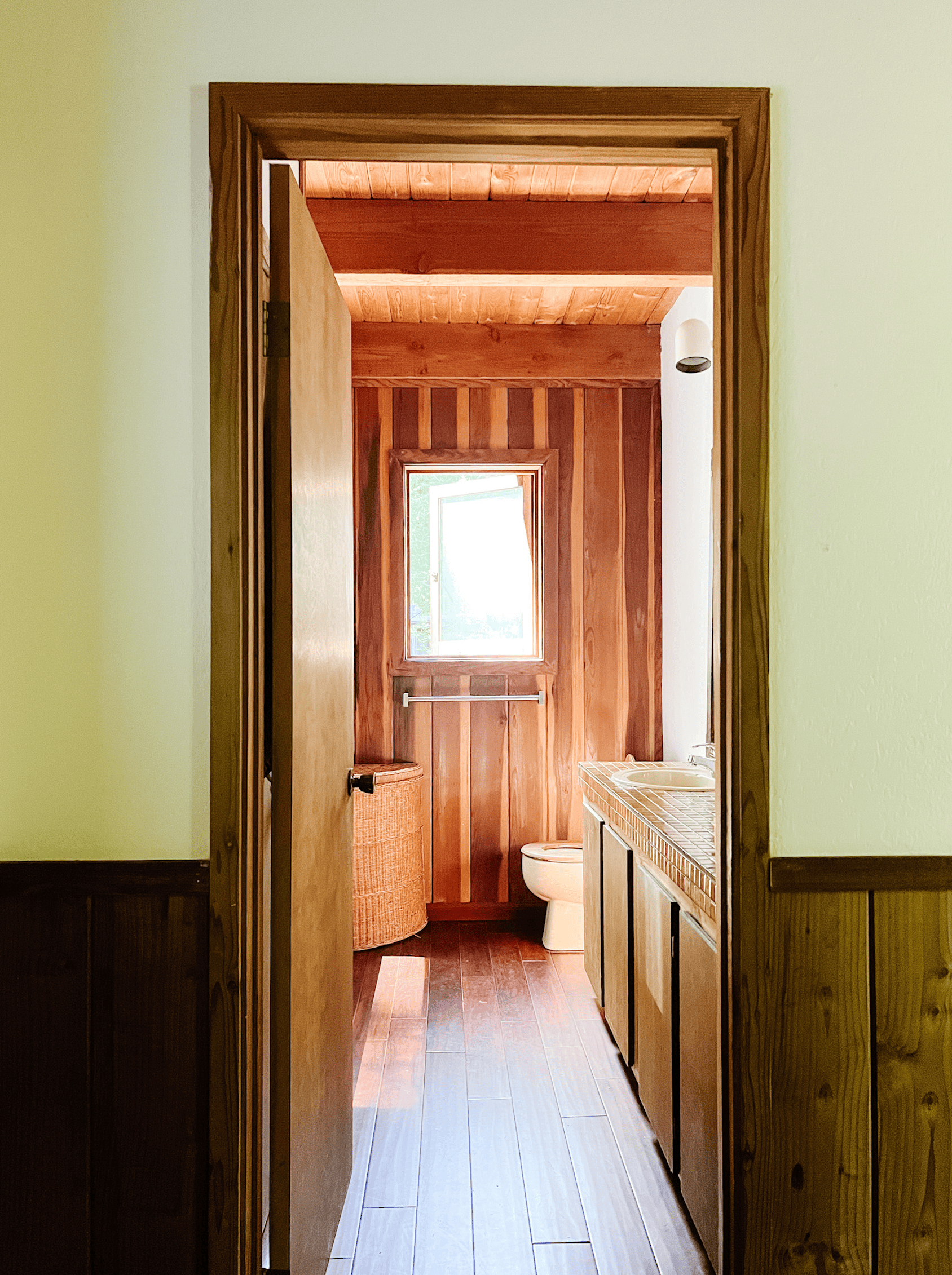 A before cabin bath features a redwood ceiling, builder-grade floors, a decaying vanity, and a picture window at the end. 