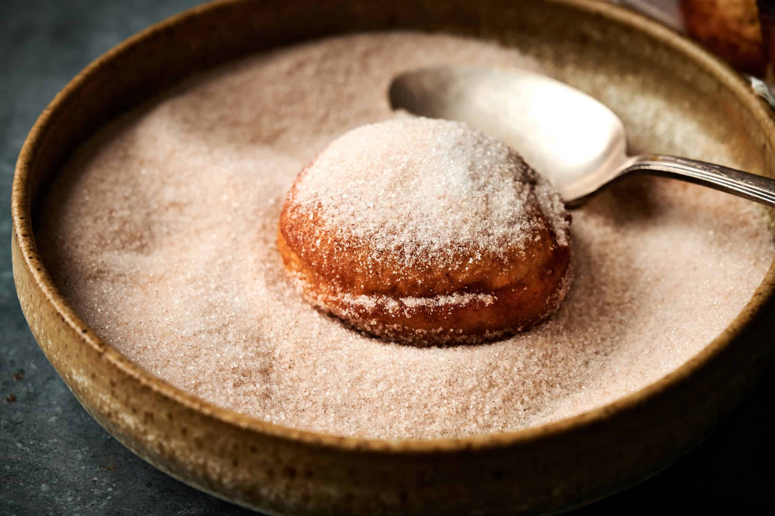 a single applesauce fritters sitting in a bed of sugar and cinnamon with a spoon. 