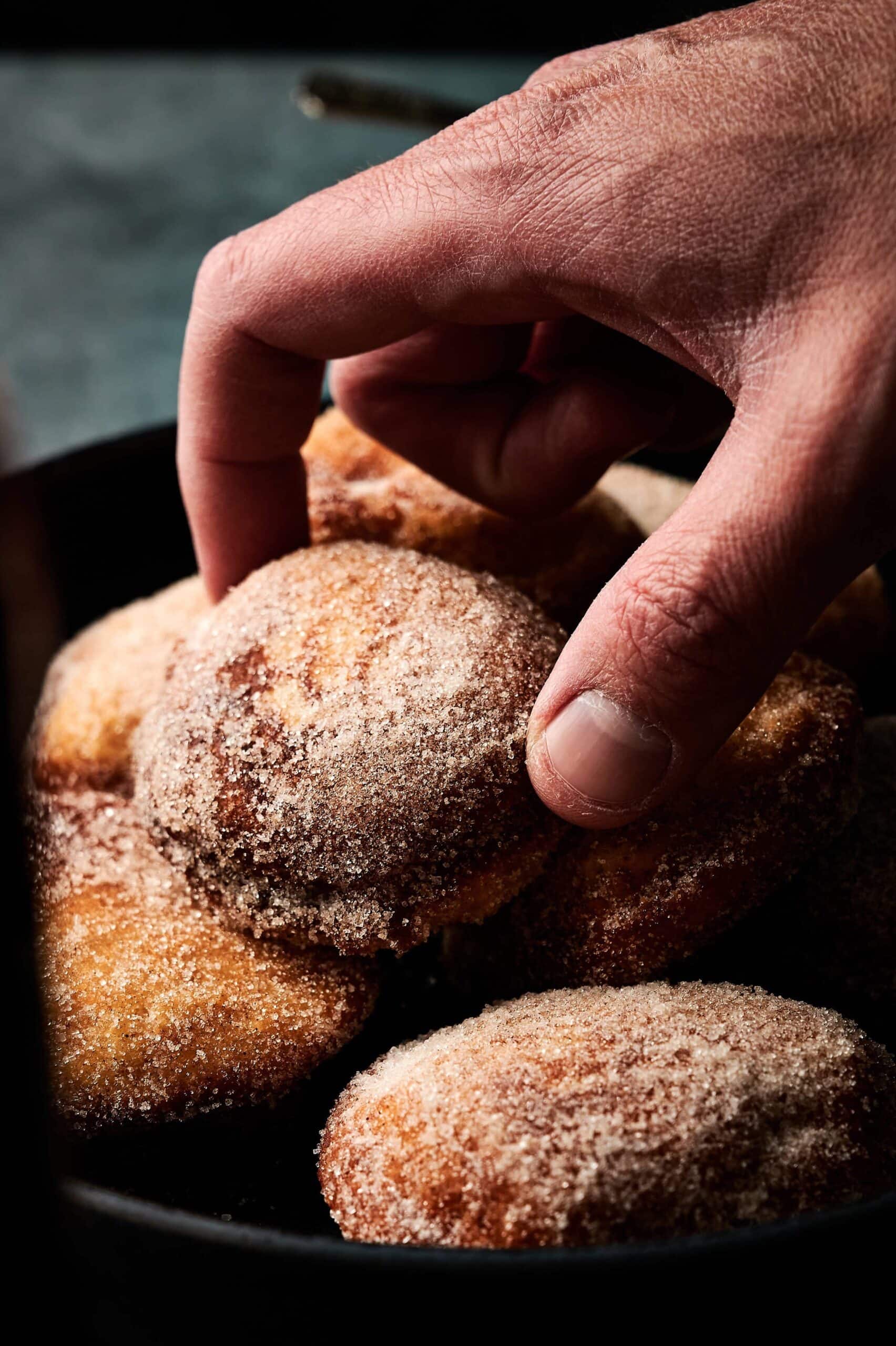Applesauce fritters dusted in sugar and cinnamon mixture up close on a black bowl with a man's hand coming down to grab one.