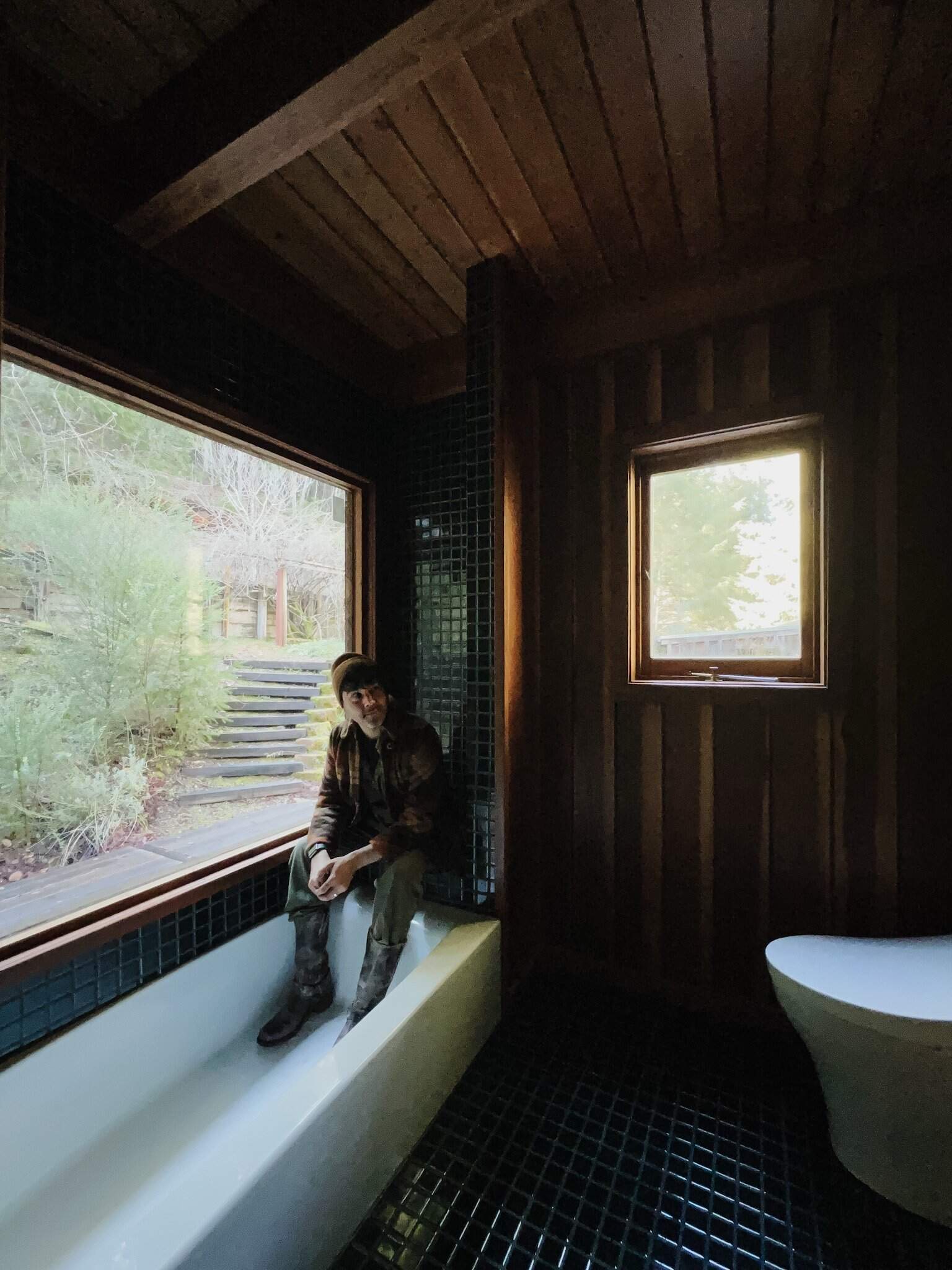 Anthony sitting in the moody bath bench in a flannel jacket admiring the shiny green 2x2 tile and refreshed redwood window. 