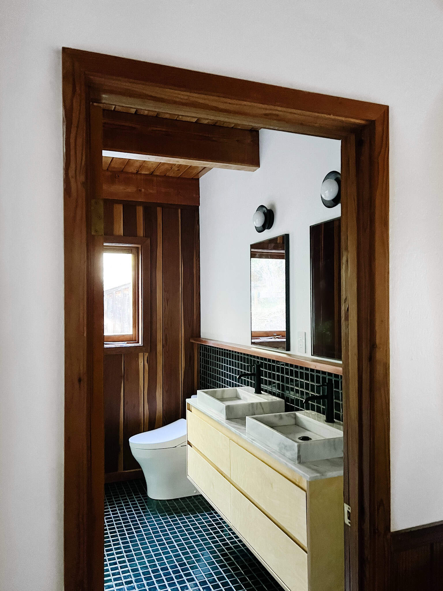 looking through a doorframe you see a renovated moody bath with brand new evergeen tile, birch vanity with modern marble sinks and new conces and windows.