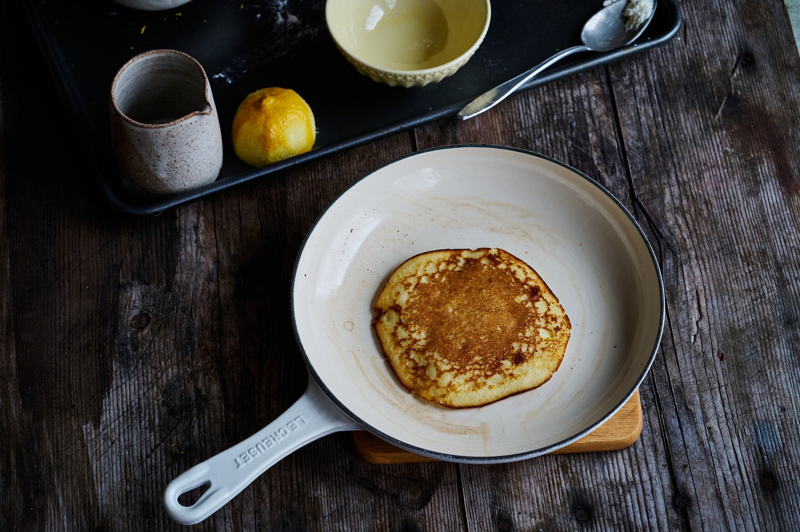 a cooked lemon and ricotta pancake golden brown in a pan with lemon and syrup in a jar on the side, on top of a wooden table.