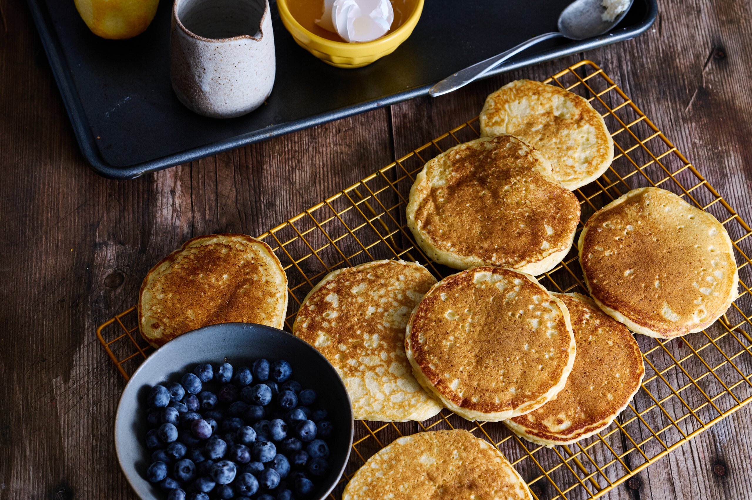 a table full of fluffy, golden-brown lemon and ricotta pancakes with blueberries cooling on a rack.