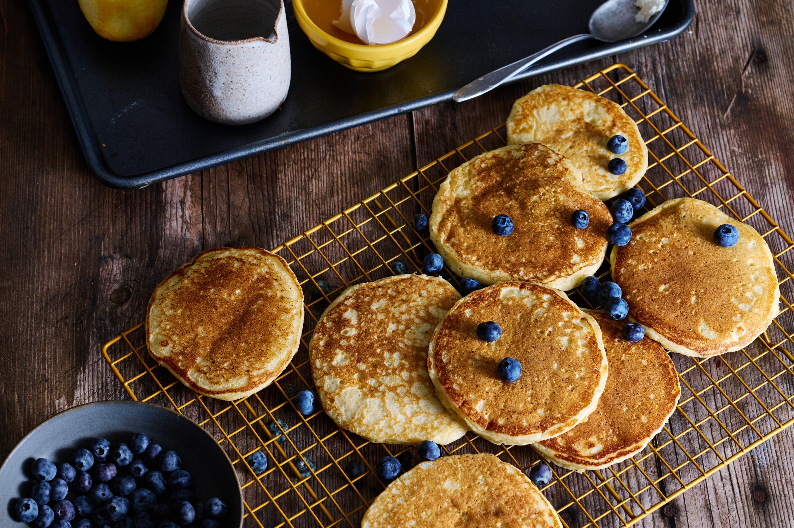 a table full of fluffy golden brown lemon and ricotta panckaes with blueberries cooling on a rack.