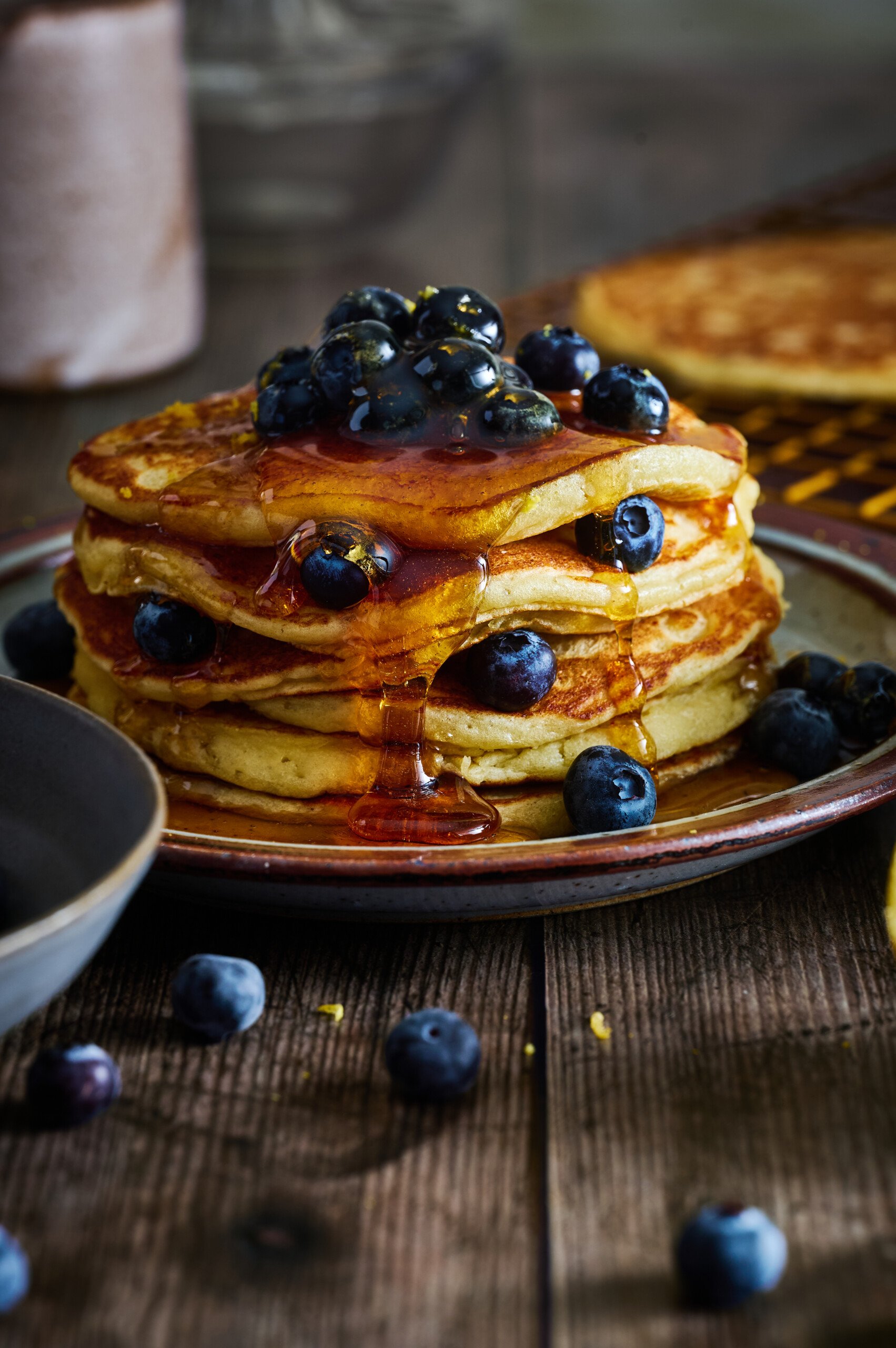 Lemon and Ricotta Pancakes iclose up looking gorgeous with glossy maple syrup and fresh blueberries stacked high on a wooden table.