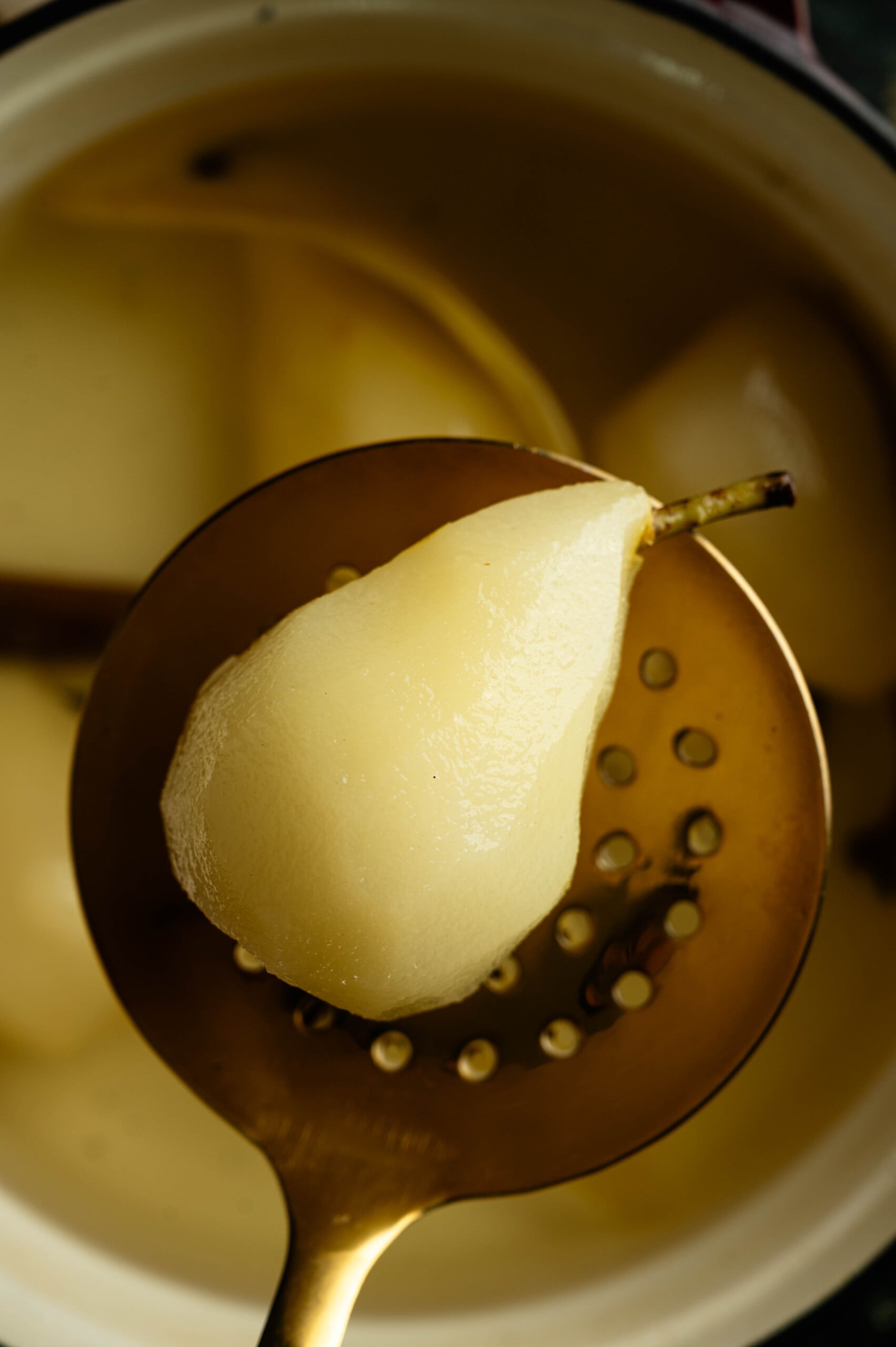 Testing a poached pear with a knife to check if the fruit is tender showing a pear above the liquid in a slotted spon,